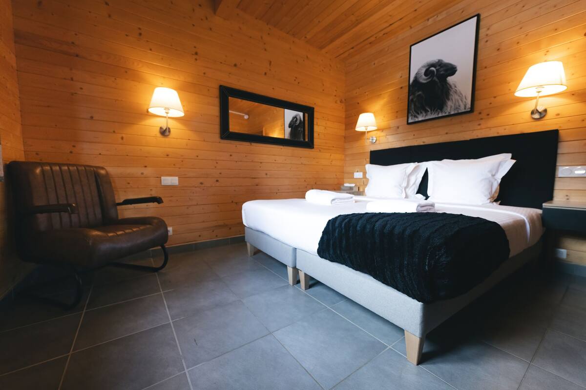 A wooden-paneled room with a bed, two wall-mounted lamps, a chair, and a framed picture on the wall.