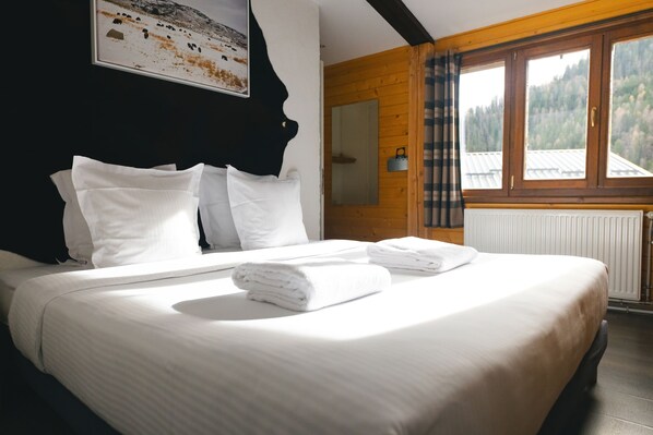 A neatly made bed with white linens and towels, a wooden headboard, a window with a view of a forest, and a framed picture on the wall. - Hôtel Le Loup Blanc (Uvernet-Fours)