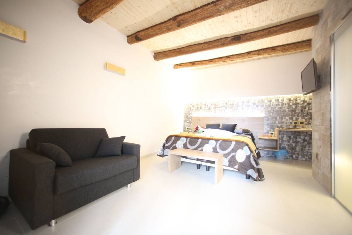 A modern bedroom with a grey sofa, a bed with a patterned bedspread, and a wooden ceiling.