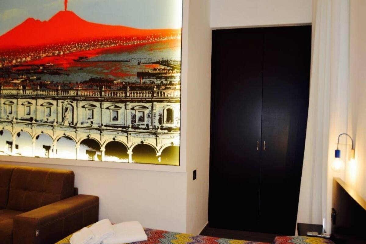 A hotel room with a bed, a brown sofa, a colorful bedspread, a large wall painting of a cityscape, and a dark door.