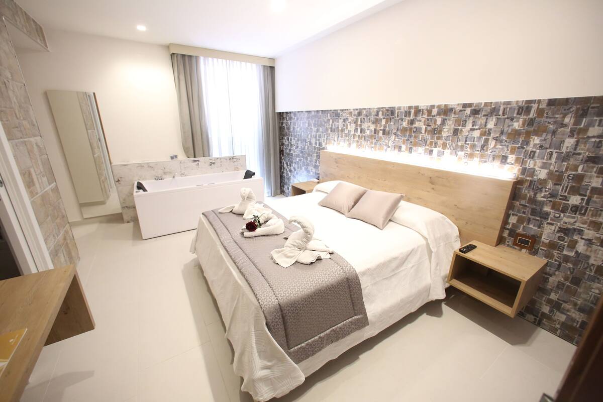 A modern hotel room with a large bed, a wooden headboard, a built-in shower, and a tiled wall.