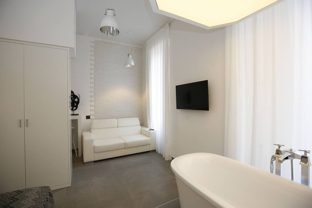 A modern bathroom with a freestanding bathtub, a white sofa, and a flat-screen TV mounted on the wall.