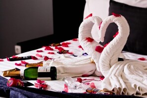 A bed with a heart-shaped towel arrangement, a bottle of champagne, and scattered rose petals.