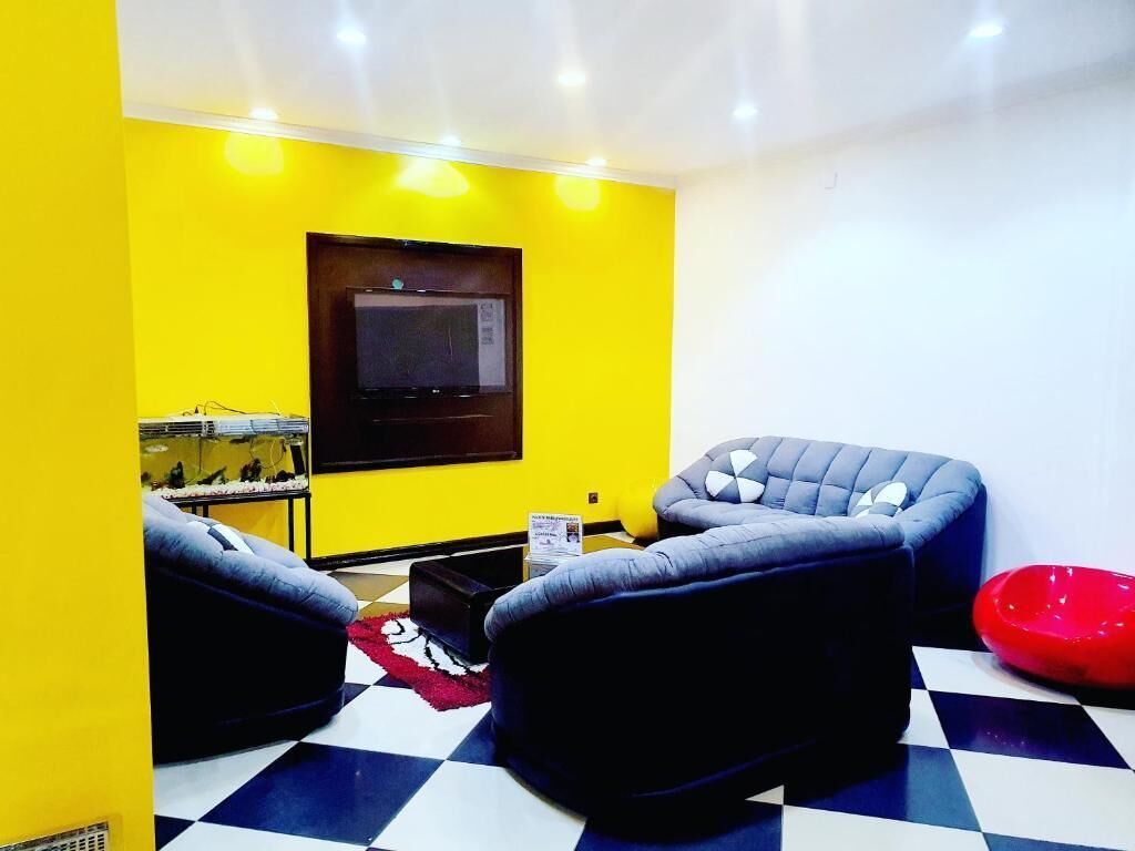 A room with a checkered floor, a flat-screen TV mounted on a yellow wall, and two black sofas.