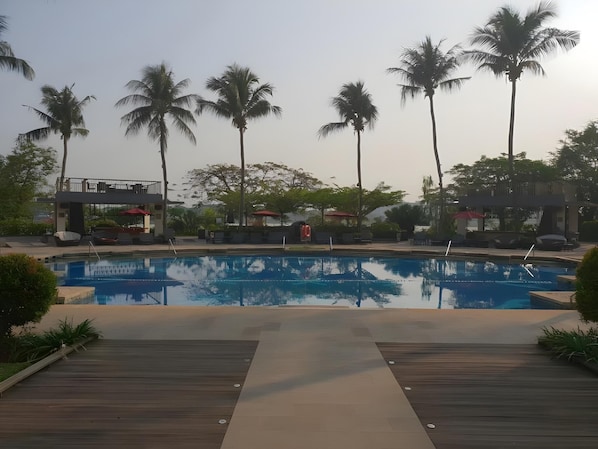 A swimming pool with a wooden deck, surrounded by palm trees and a covered seating area.