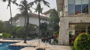 Una piscina con pared de piedra y palmeras, un patio con mobiliario exterior, y un edificio con ventanas amplias y un balcón.