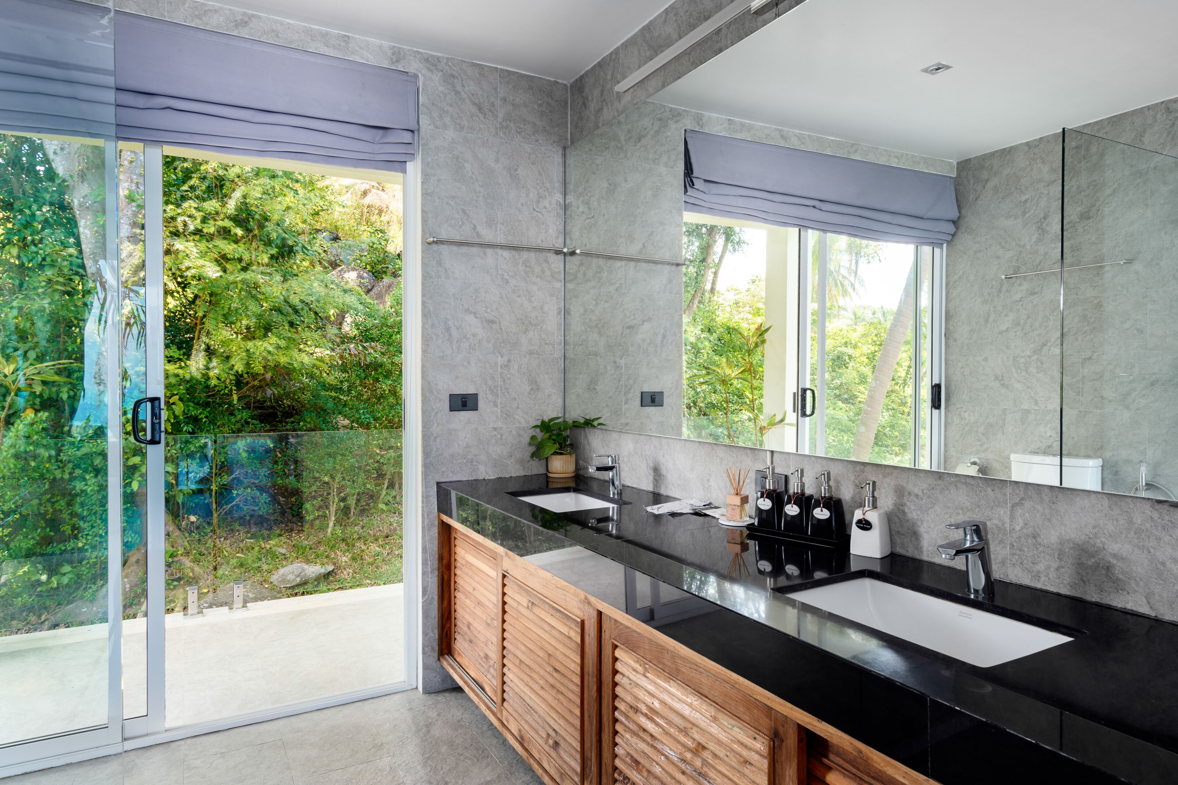 Un baño moderno con un espejo grande, dos lavamanos y un vidrio que da a un balcón con vistas al verdor.