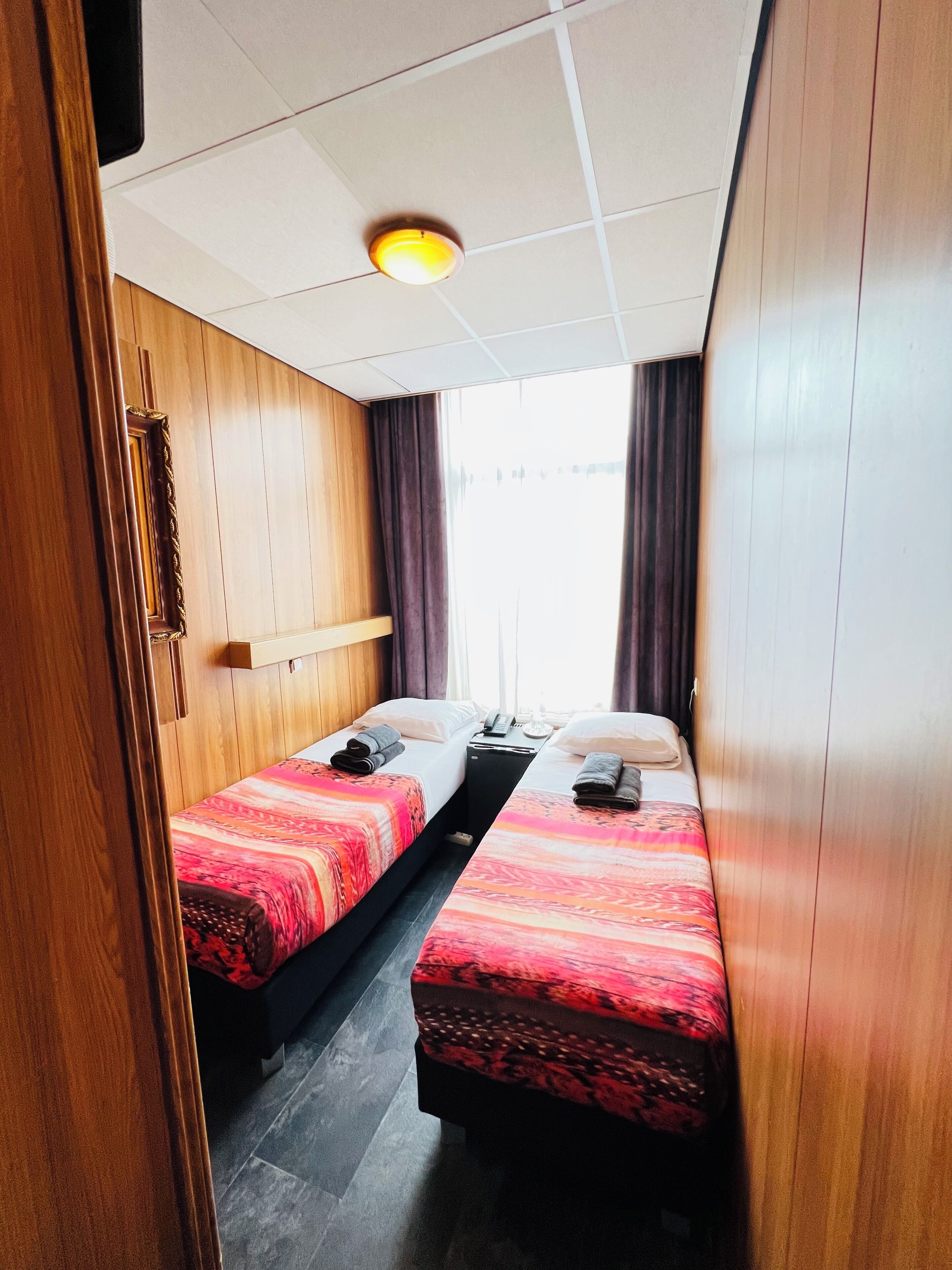 A compact hotel room with two single beds, wooden paneling, a ceiling light, and a window with curtains.