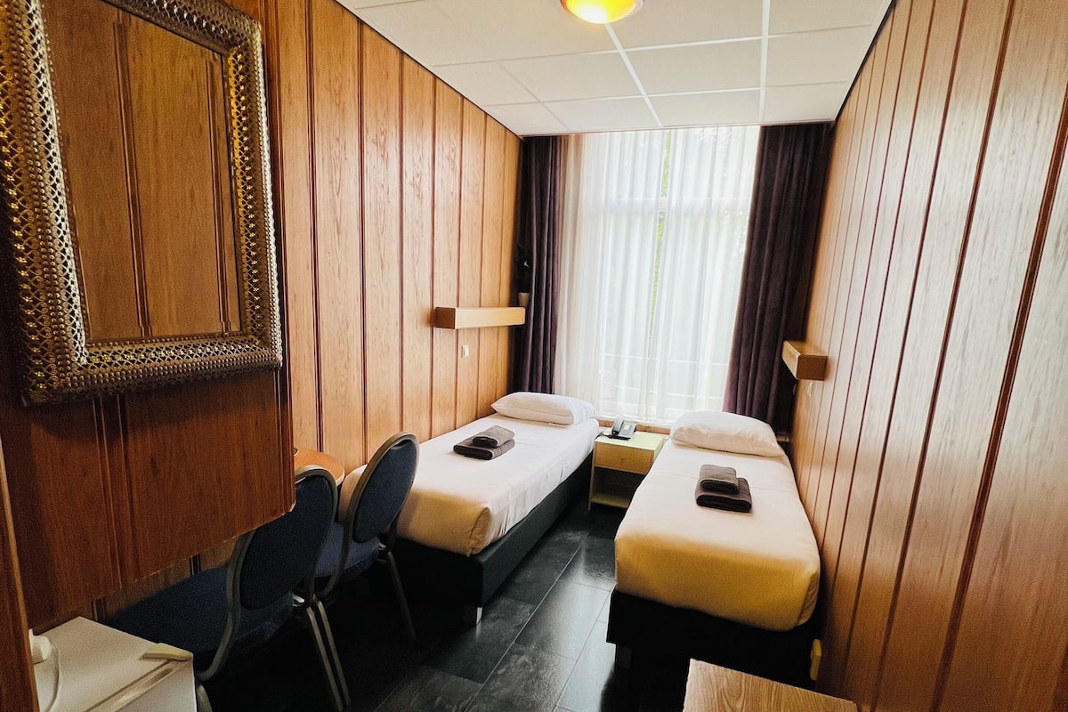 Habitación de hotel compacta con dos camas individuales, un armario de madera, una mesita, una silla y un televisor montado en la pared.