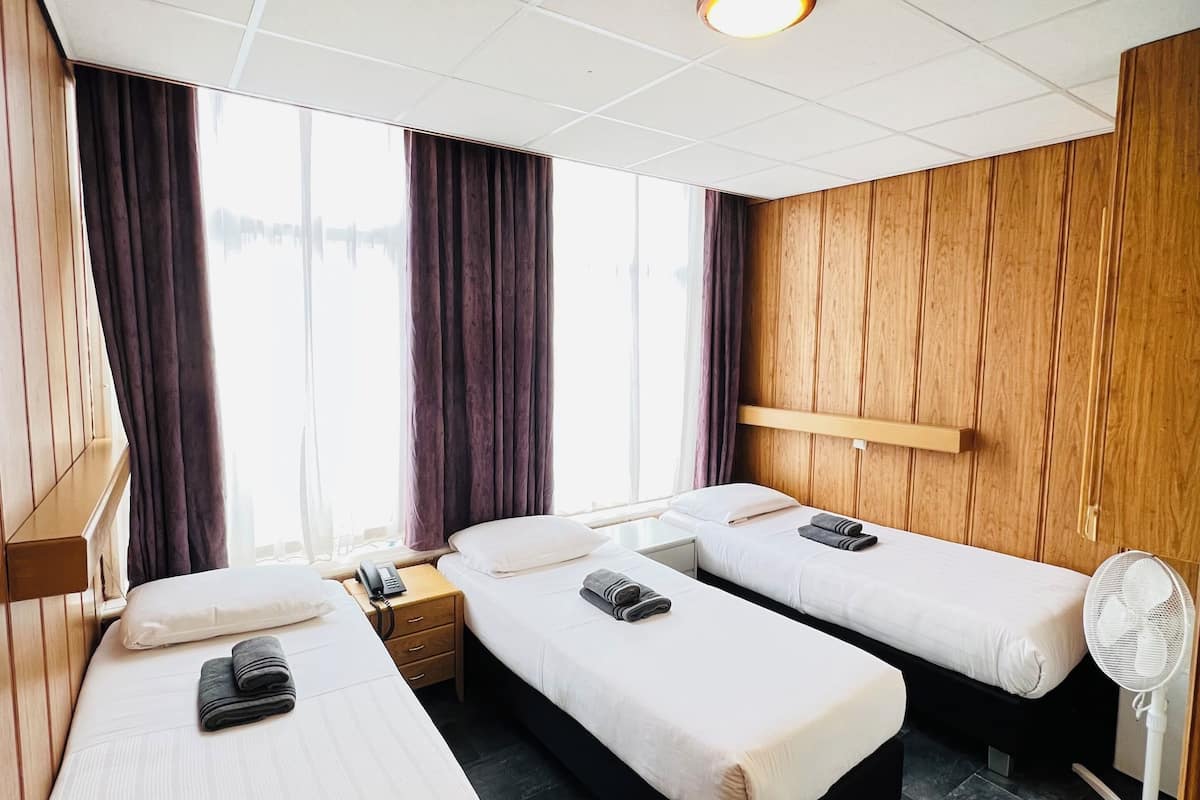 Habitación de hotel con dos camas individuales, cabecero de madera, mesita de noche con teléfono, pequeña mesa con ventilador y ventana con cortinas.