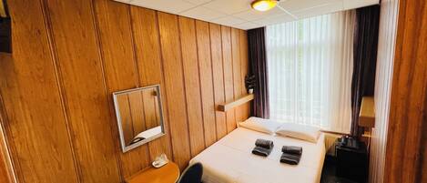 A hotel room with a wooden paneled wall, a bed with two pillows and two folded towels, a small table with a chair, a bedside table, and a window with curtains.