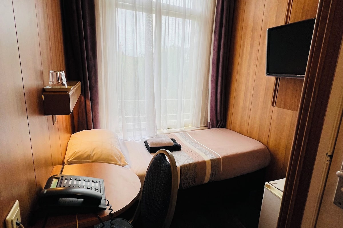 Habitación de hotel pequeña con una cama individual, un escritorio con teléfono, una silla, un televisor pequeño y una ventana con cortinas.