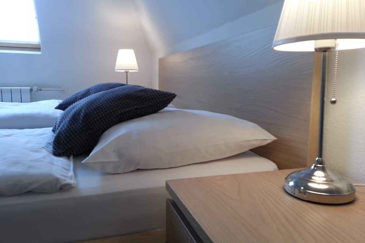 A neatly made bed with white and dark blue pillows, a wooden bedside table with a lamp, and a window with blinds in a hotel room.