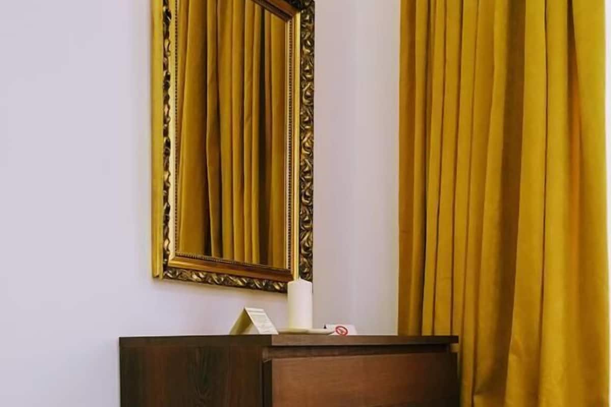 A hotel room with a wooden dresser, a bed with a white and blue pillow, a mirror with a gold frame, and yellow curtains.