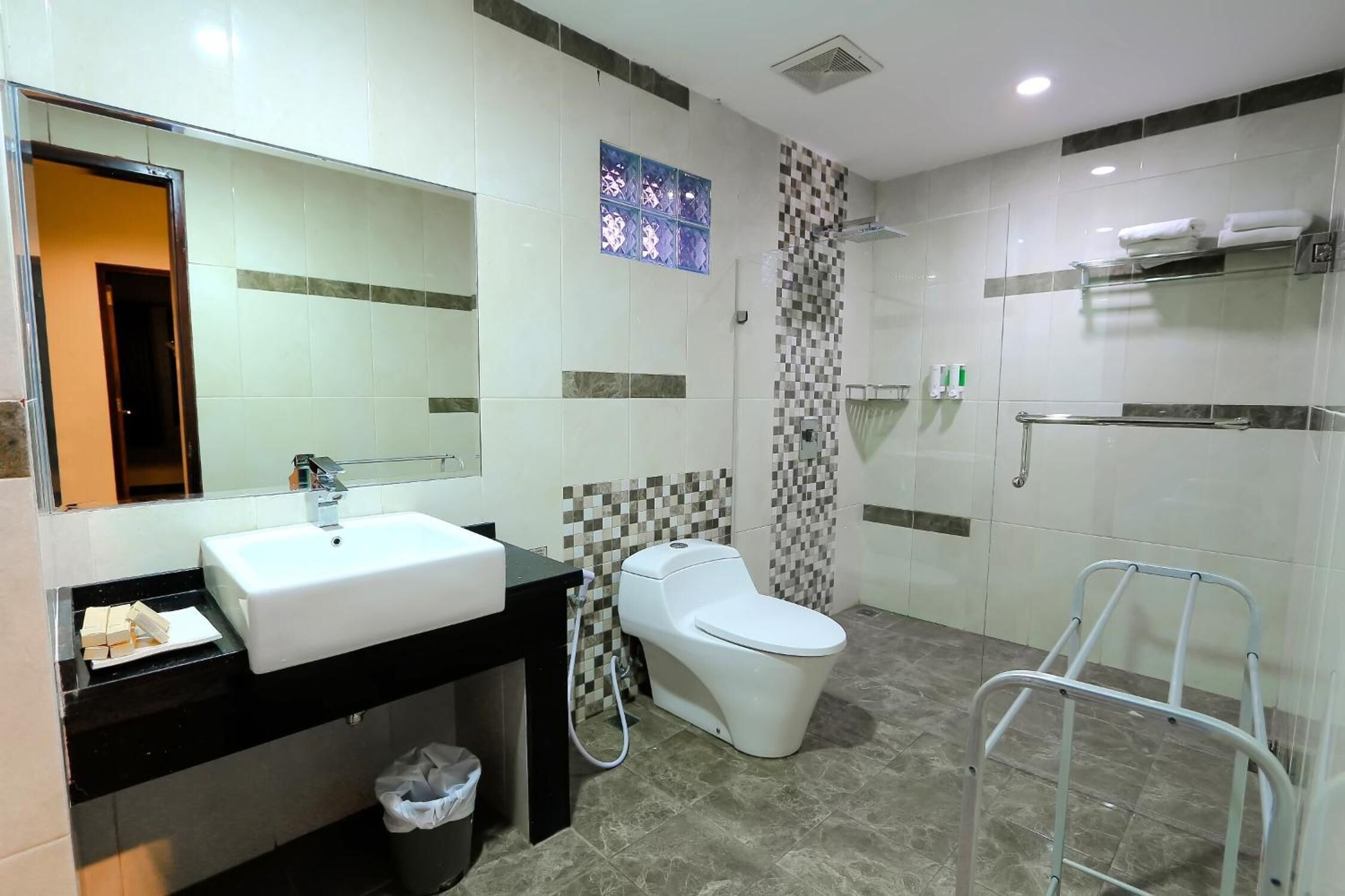 A modern bathroom with a white sink, toilet, and shower. There is a towel rack and a trash bin.