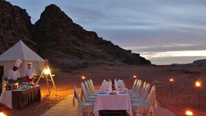 Un montaje para cenar en el desierto con una carpa, una mesa dispuesta para varios comensales y cocineros preparando comida.