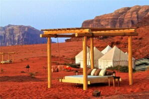 Un campement dans le désert, avec un lit-hamac en bois, des tentes et une vue sur des formations rocheuses rouges.