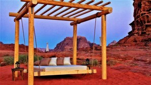 Una hamaca de madera con almohadones en un paisaje desértico con formaciones rocosas rojas al fondo.