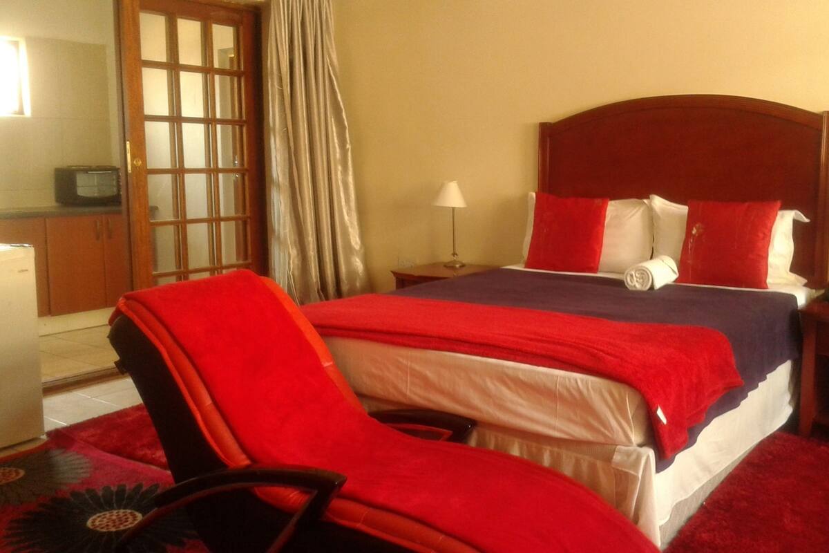 Une chambre d’hôtel comprenant un lit, une chaise, une table de chevet, une lampe, un téléviseur et une fenêtre avec des rideaux.