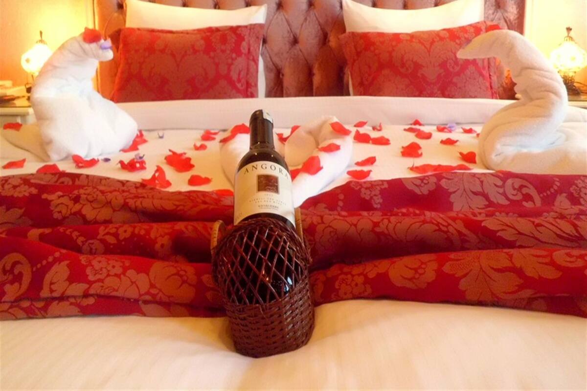 Een bed met een rode bloemenlaken, twee witte kussen in de vorm van zwanen en een fles Angora parfum.