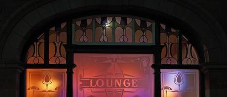 Un bar lounge con un cartel que dice "LOUNGE BAR" bajo un ventanal arqueado con vidrieras decorativas.