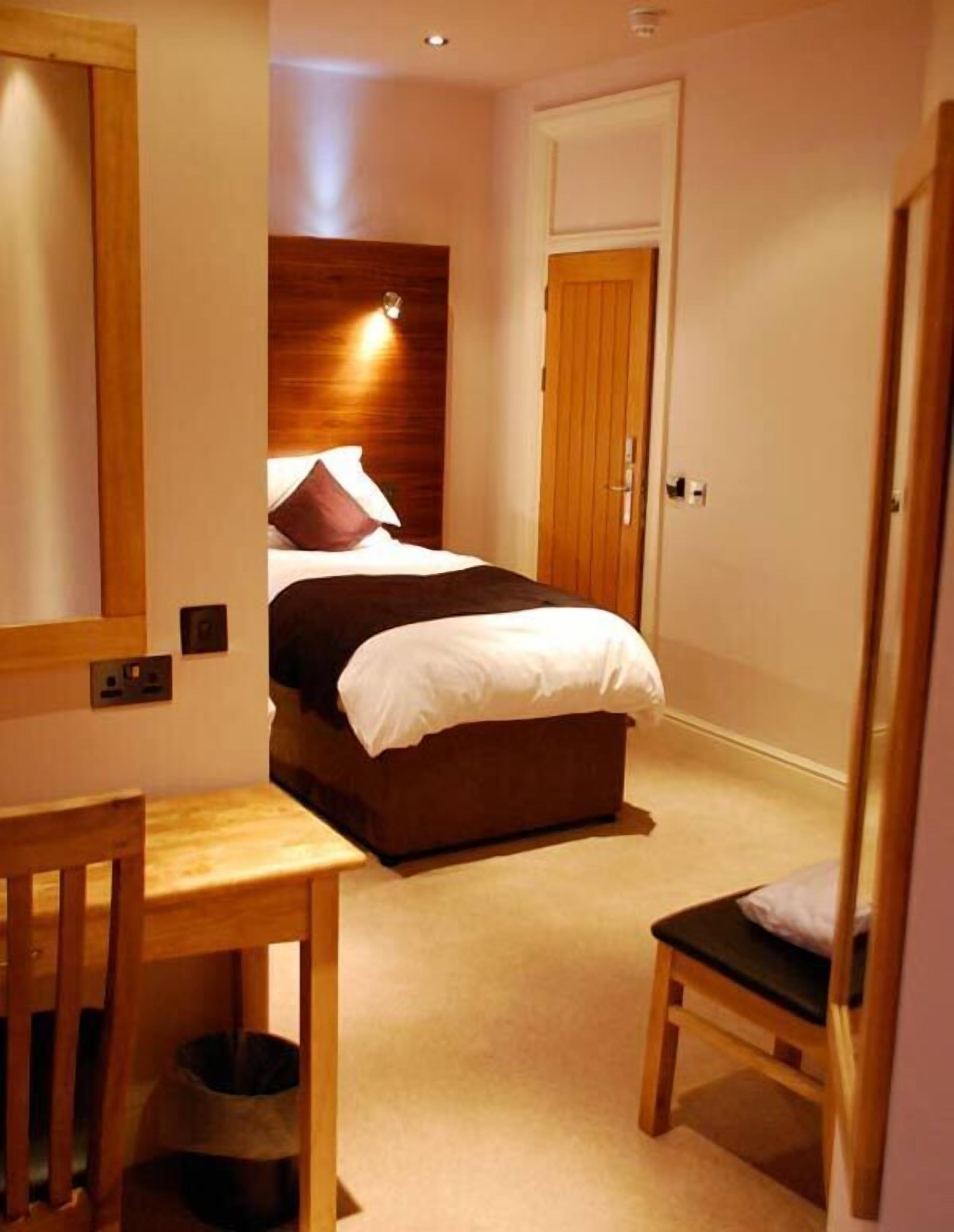 Een slaapkamer met een bed, nachtkastje, stoel en een kleine prullenbak.