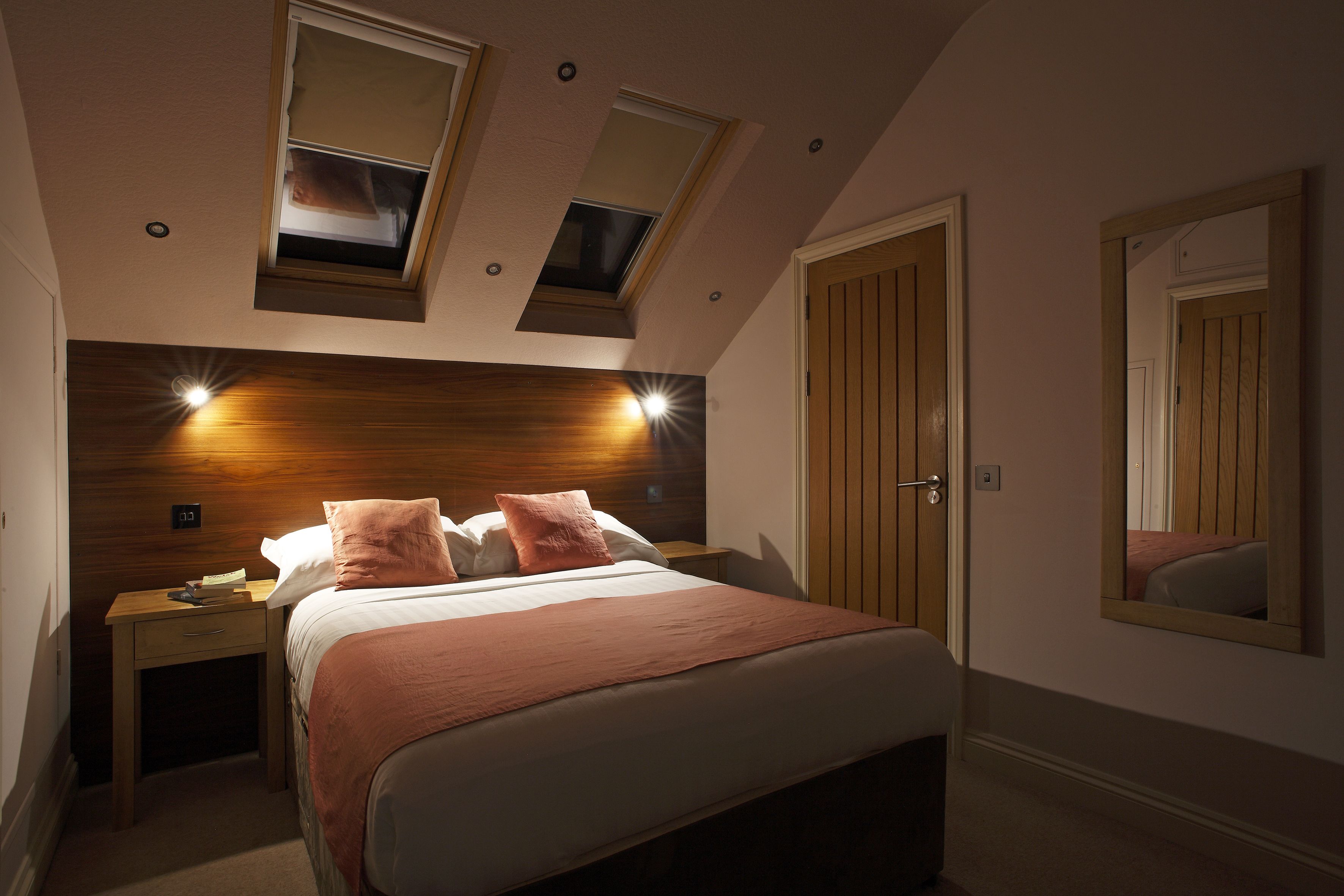 Un dormitorio con cama, dos mesitas de noche, una mesita auxiliar, cabecera de madera, dos tragaluces, un espejo y una puerta.