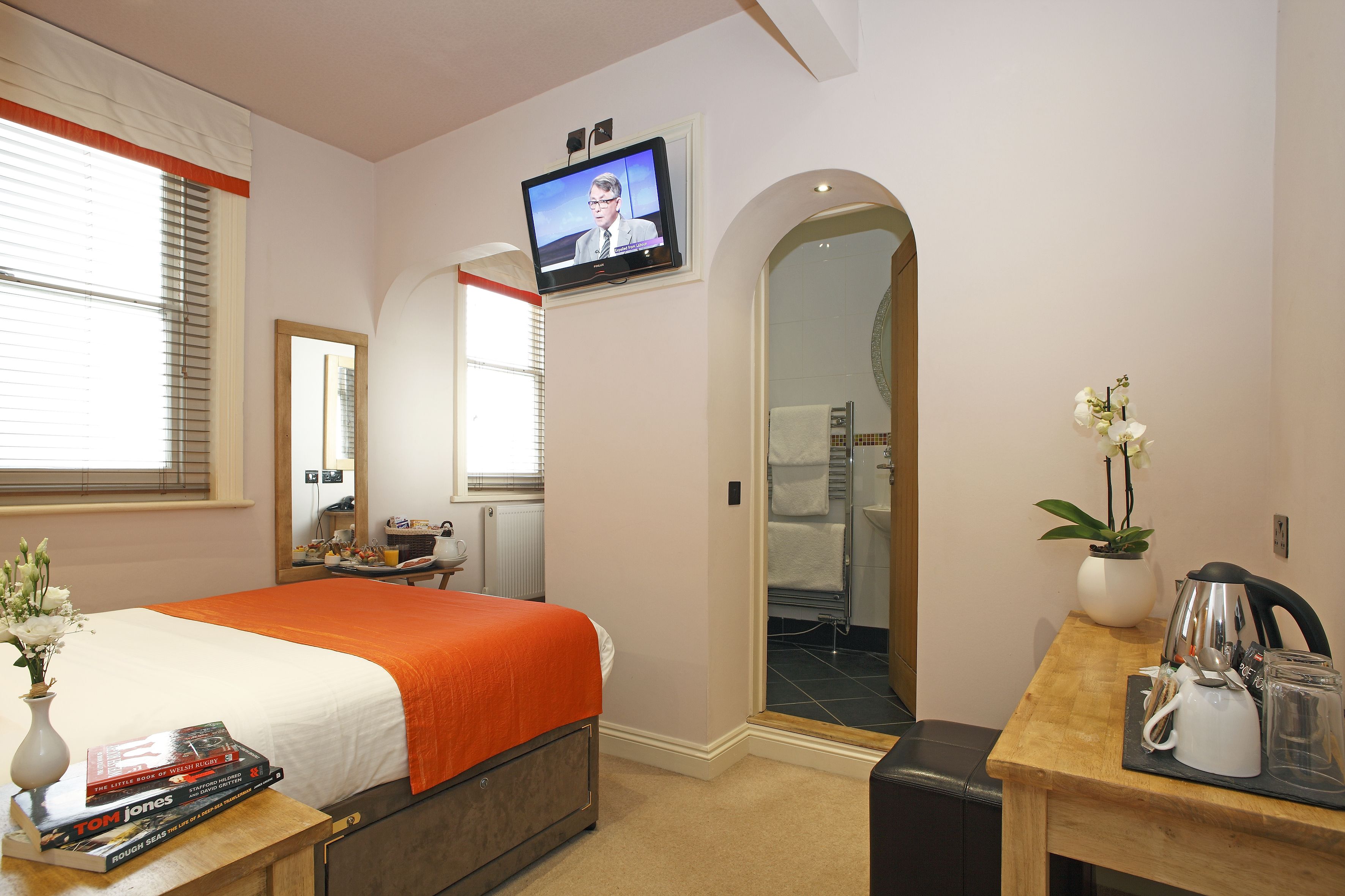 Habitación de hotel con cama, televisor montado en la pared, escritorio con tetera, un florero con flores y un espejo.