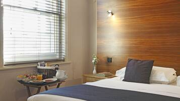 A hotel room with a wooden headboard, a bed with white linens, a bedside table with a basket, a vase with flowers, and a breakfast tray.