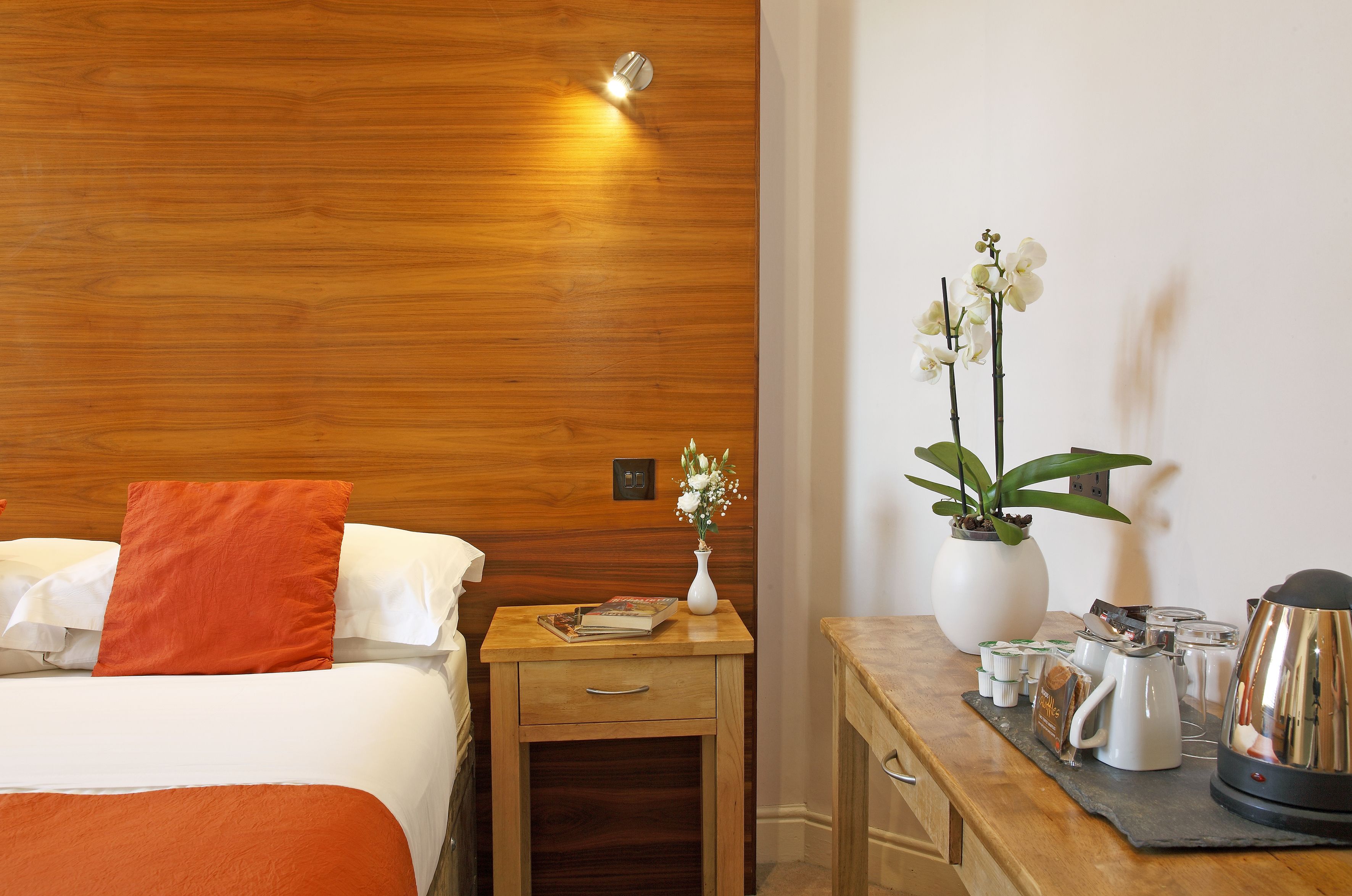 A hotel room with a wooden headboard, a bed with white and orange bedding, a nightstand with a vase of flowers, a coffee station, and a small table with a book.