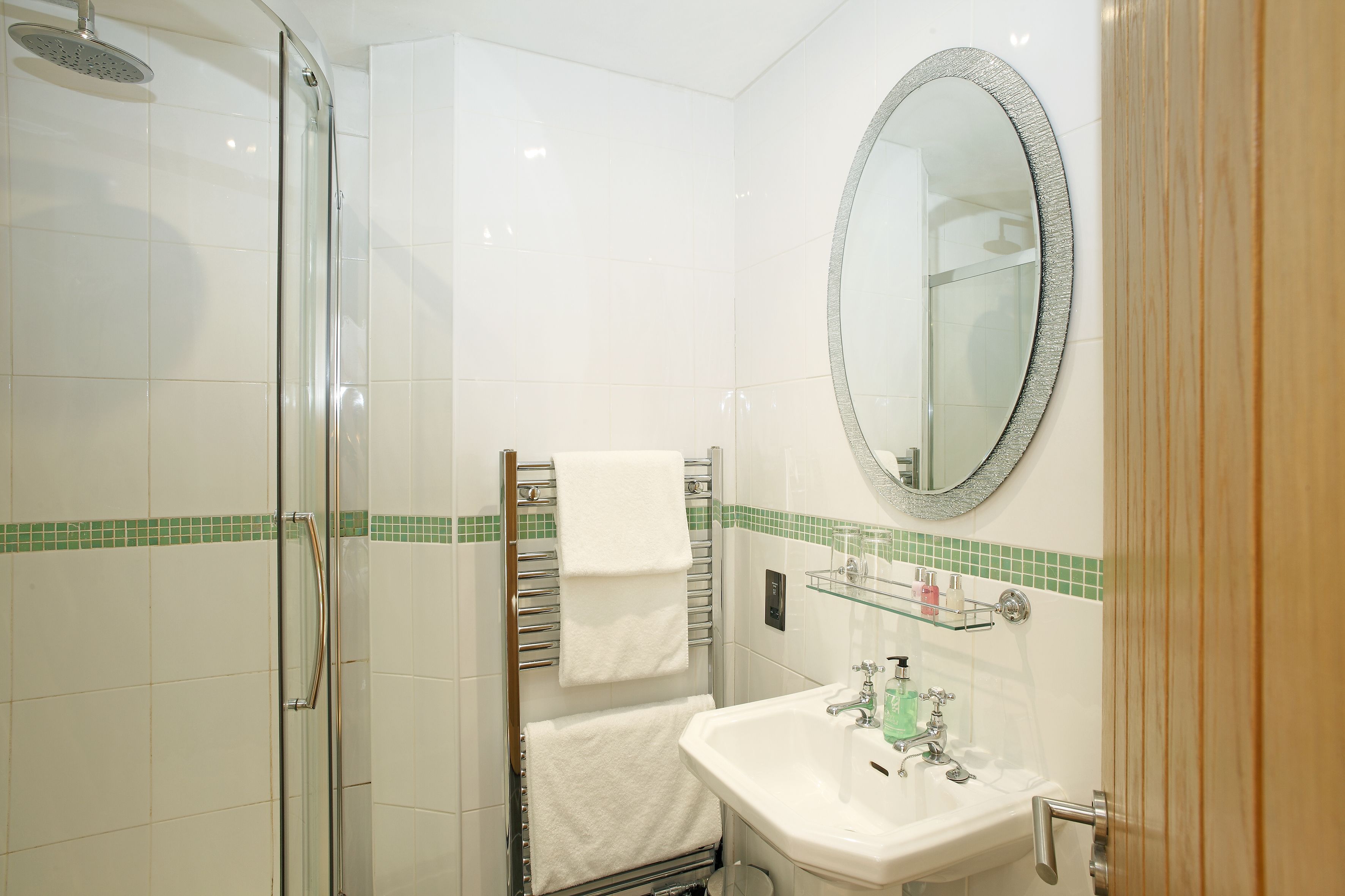A bathroom with a glass shower enclosure, a white sink with a green soap dispenser, and a mirror with a decorative frame.