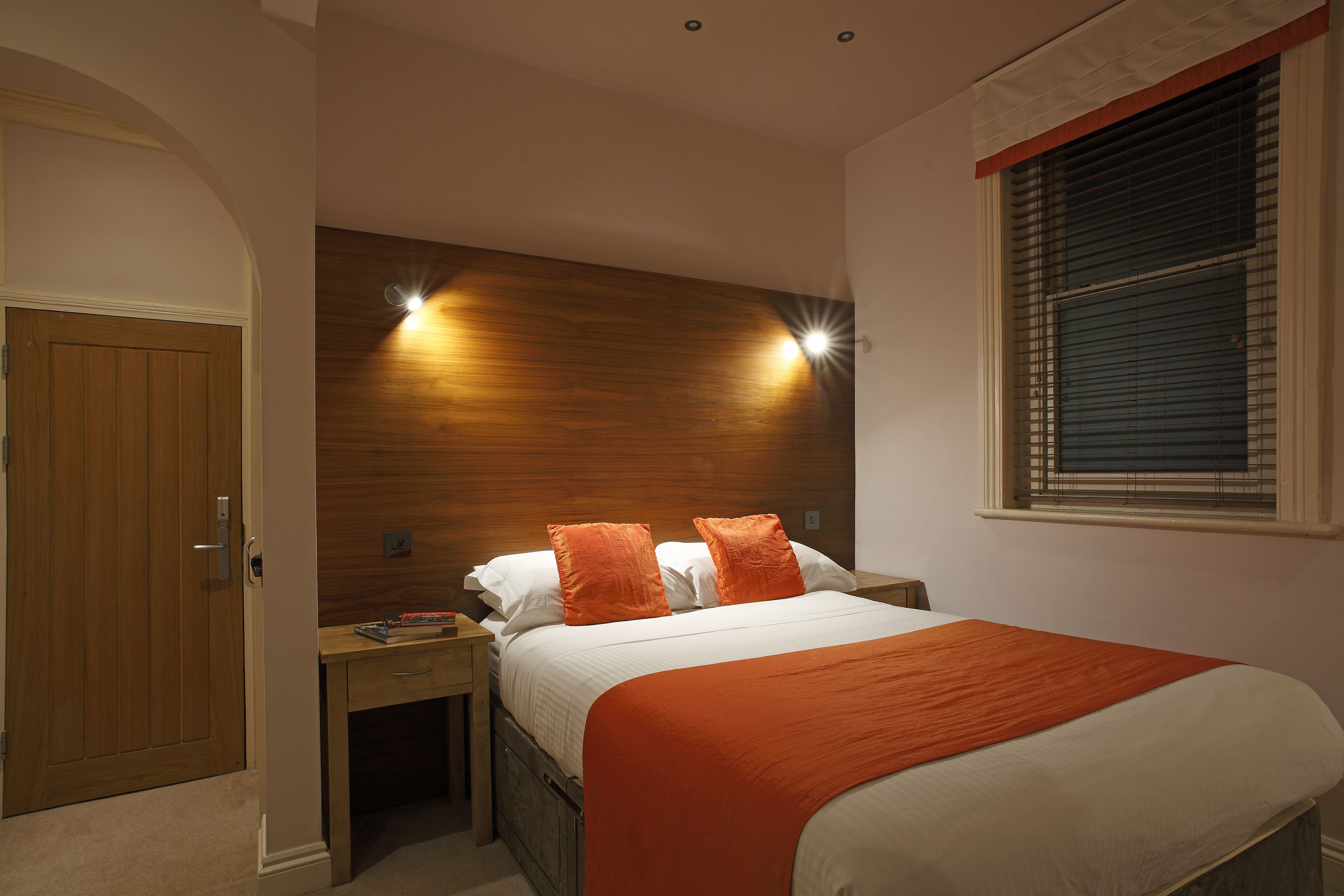 Un dormitorio con cabecera de madera, dos mesitas de noche, una cama con ropa de cama blanca y naranja, y una ventana con persianas.