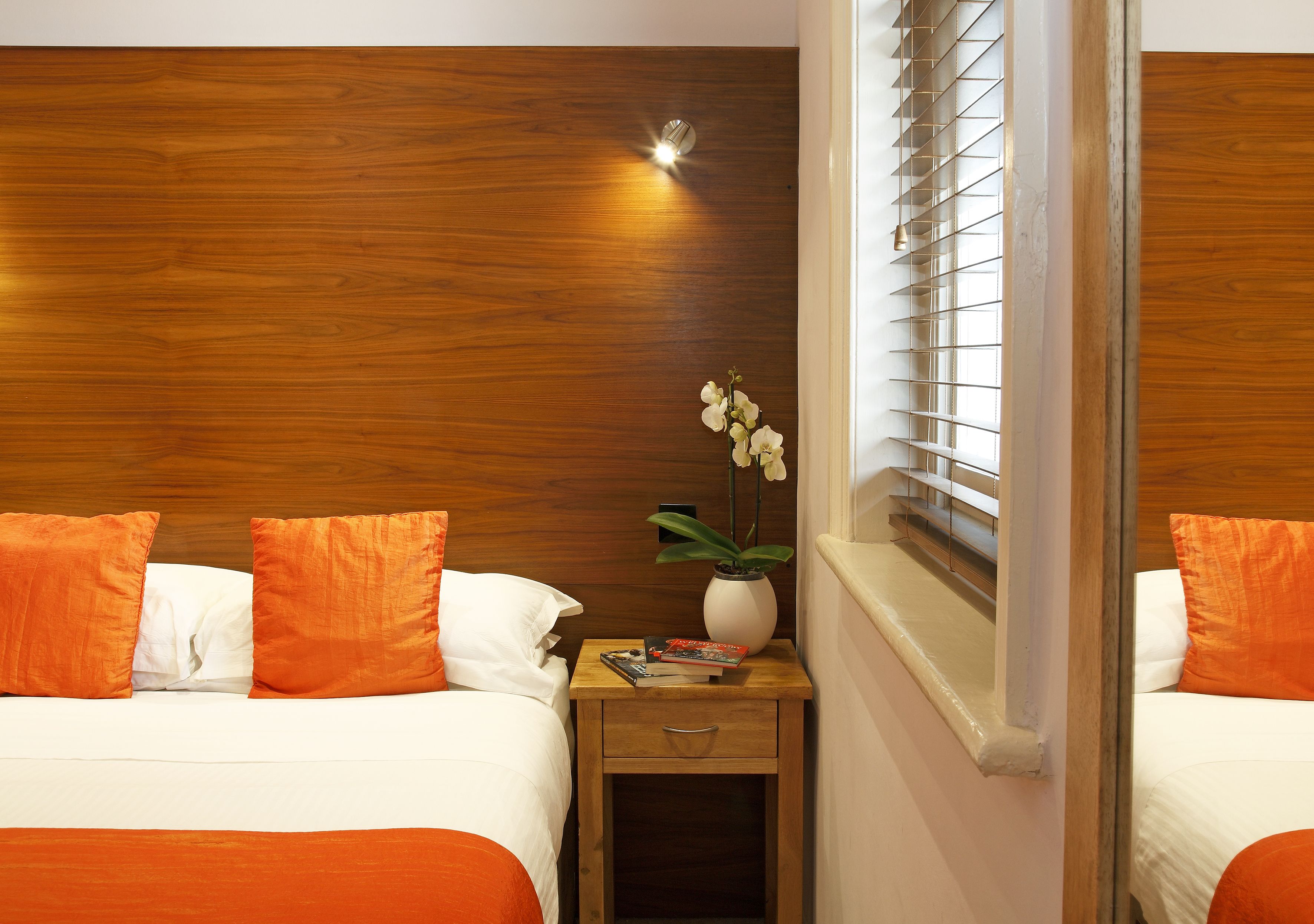 A hotel room with two beds, wooden headboard, a nightstand with a vase of flowers, and a window with blinds.