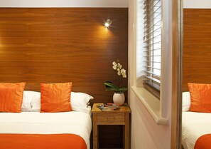 A hotel room with two beds, wooden headboard, a nightstand with a vase of flowers, and a window with blinds.