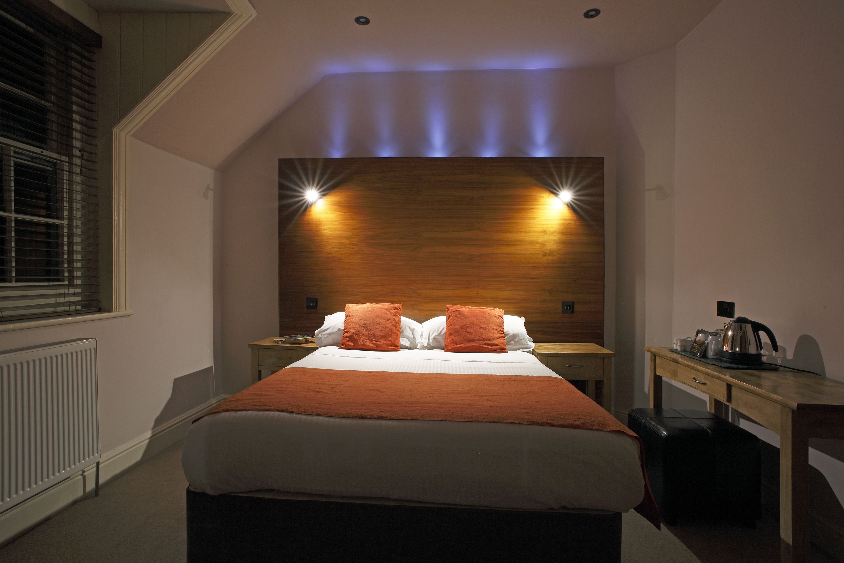 Habitación de hotel con una cama grande, dos mesitas de noche, un cabecero de madera con iluminación integrada, un radiador, un escritorio y una silla.