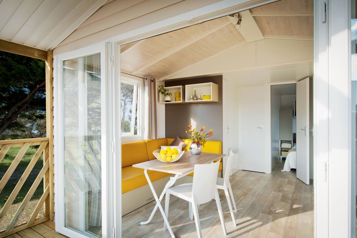 A modern, open-plan living area with a wooden deck, a yellow sofa, a dining table with chairs, and a kitchen area visible through an open door.