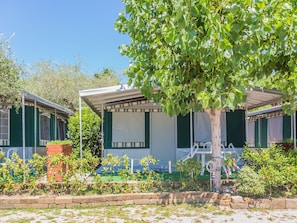 A row of green-roofed cabins with white walls and large windows, surrounded by a garden with various plants and a tree. - Camping Porto (Moniga del Garda)