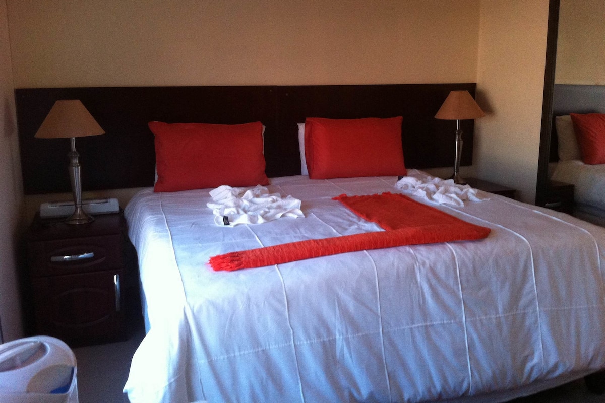 A neatly made bed with white linens, two red pillows, and a red throw blanket. There are two bedside lamps on either side of the bed.