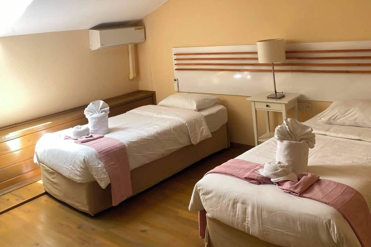 A hotel room with two beds, a skylight, a bedside table with a lamp, and a wall-mounted air conditioning unit.