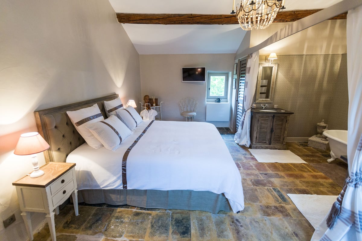 Une chambre avec un grand lit, une table de chevet avec une lampe, un téléviseur fixé au mur, une fenêtre avec des rideaux et une salle de bain avec une baignoire et un lavabo.