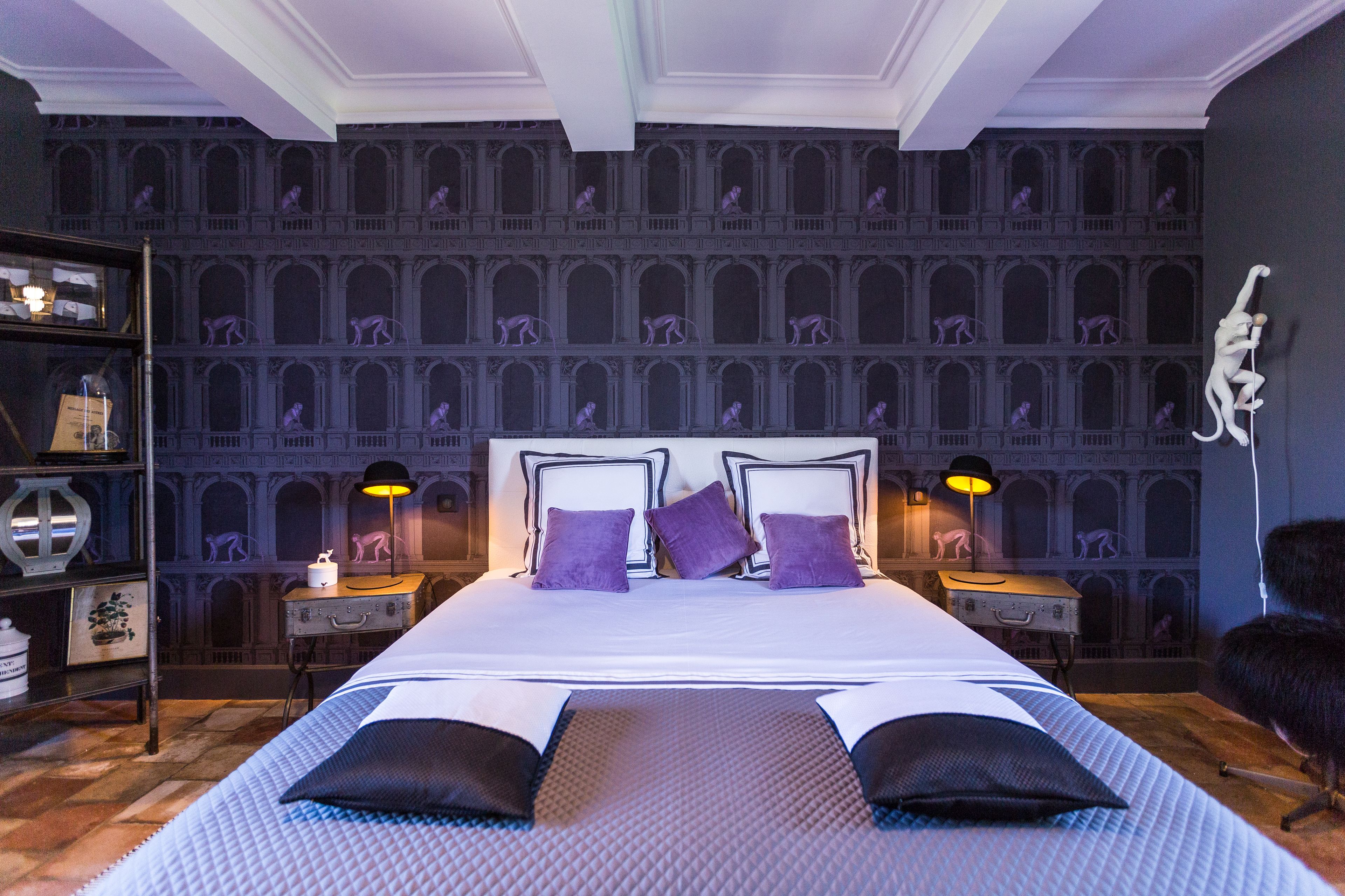 A large bed with a headboard, two bedside tables with lamps, a bookshelf, and a decorative wall with arched windows.
