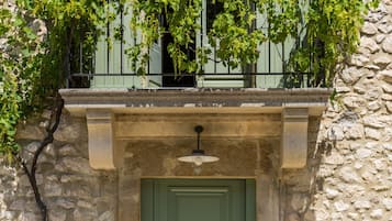 A stone building with a green door and shutters, a balcony with vines, and a small table with chairs outside.