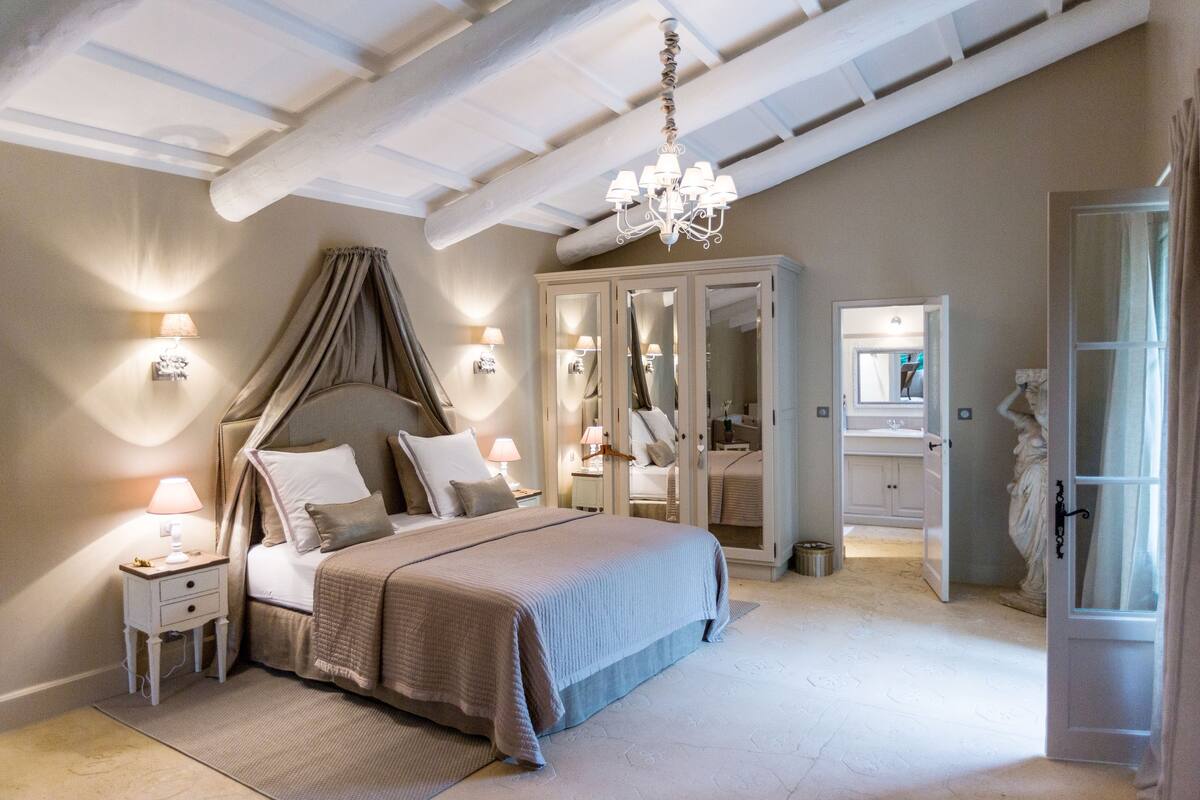 Une chambre à coucher avec un lit à baldaquin, des lampes de chevet, un lustre et un dressing doté d’un miroir.