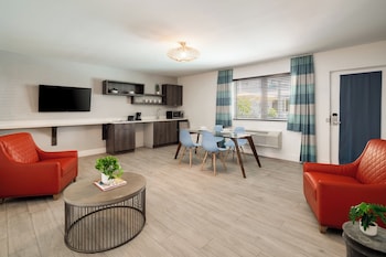 A modern living room with a flat-screen TV, two black chairs, a glass table, a grey sofa, and a framed artwork on the wall. at Waterside Hotel and Suites