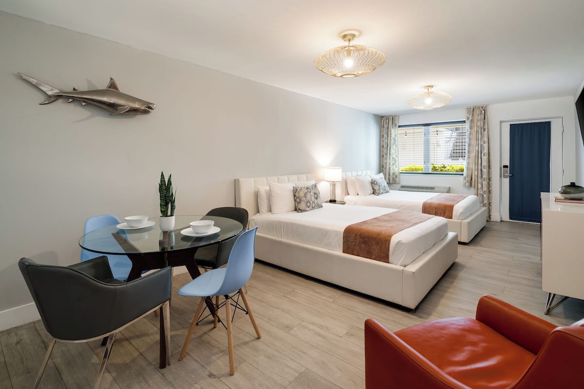 A modern hotel room with a glass dining table, two beds, a desk, and a large window with curtains.