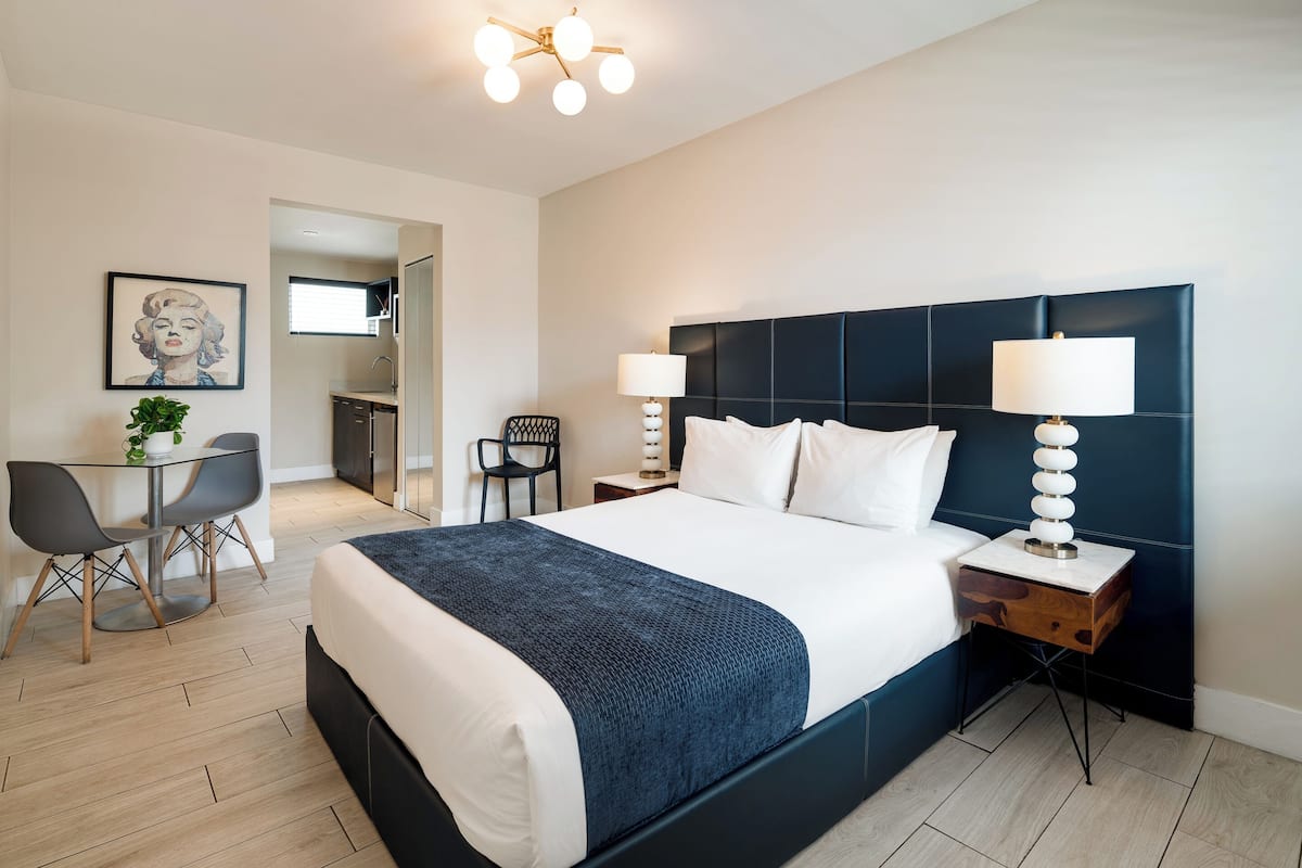 A modern hotel room with a large bed, two bedside tables with lamps, a desk with a chair, and a small table with a plant.