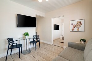 Una sala de estar moderna con un televisor de pantalla plana, dos sillas negras, una mesa de vidrio, un sofá gris y una obra de arte enmarcada en la pared.