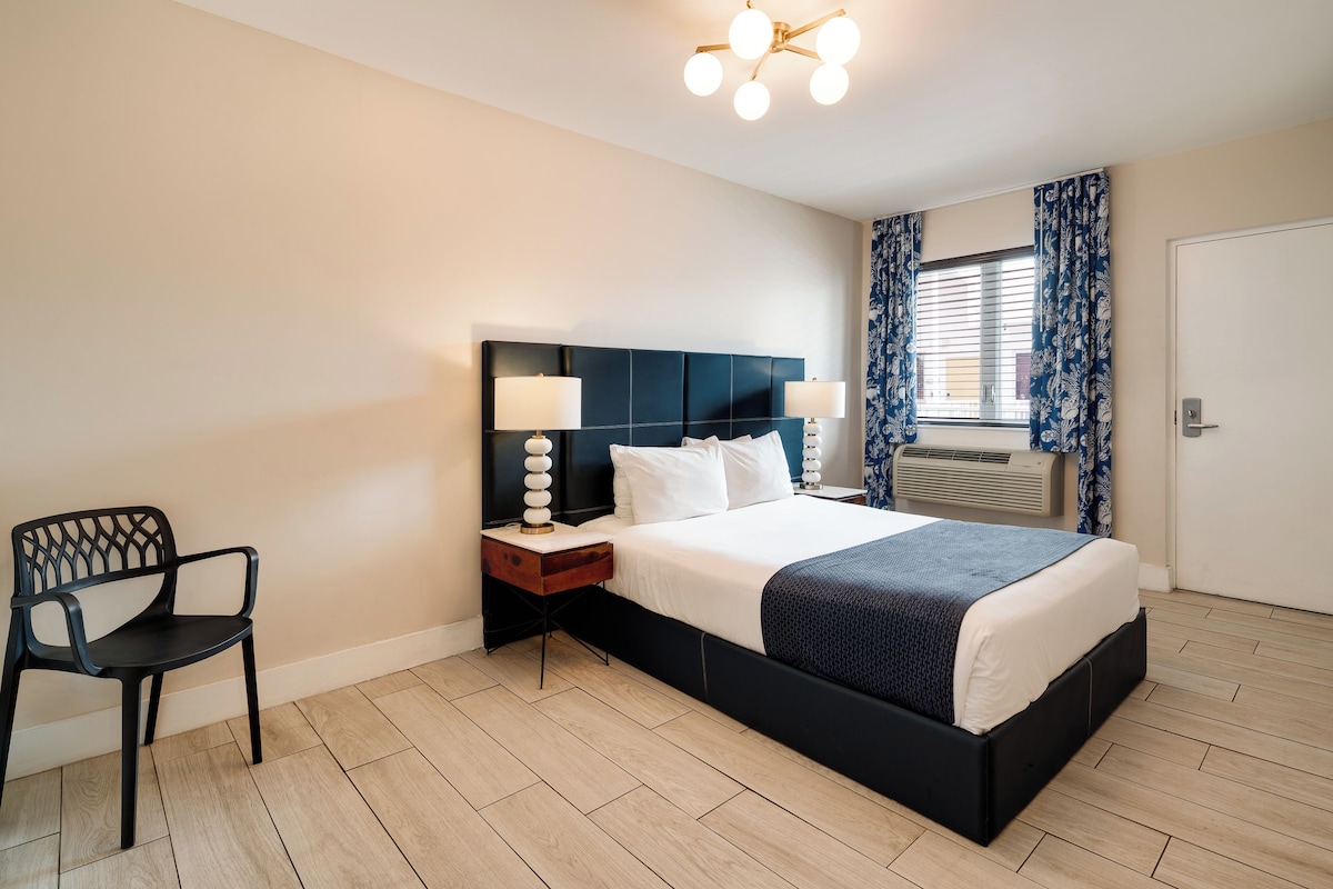 A modern hotel room with a large bed, a nightstand with a lamp, a chair, and a window with curtains.