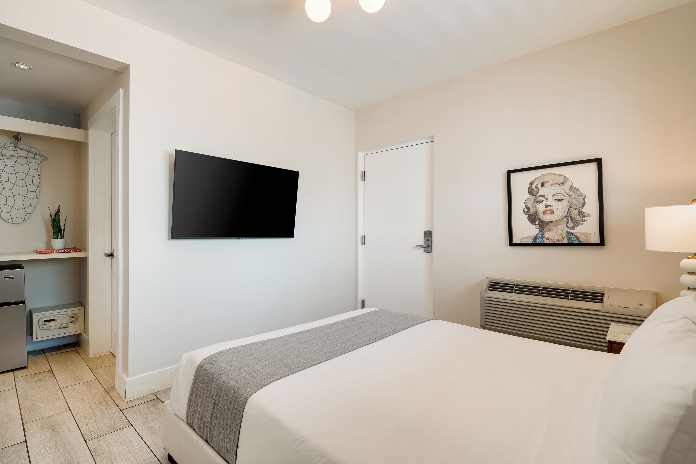 Habitación de hotel con una cama, televisor de pantalla plana montado en la pared, un cuadro enmarcado, un calentador y una pequeña cocina con refrigerador y microondas.