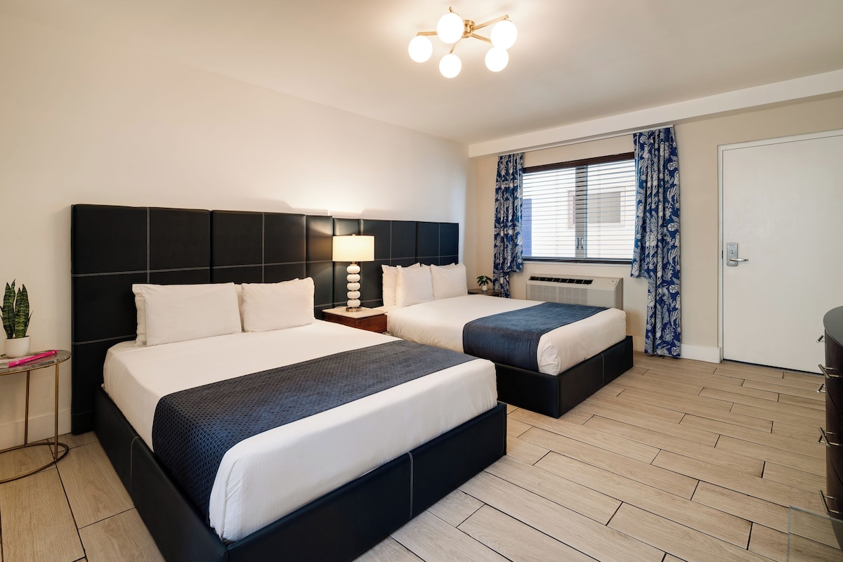 A double room with two beds, a bedside table with a lamp, a window with curtains, and a door.